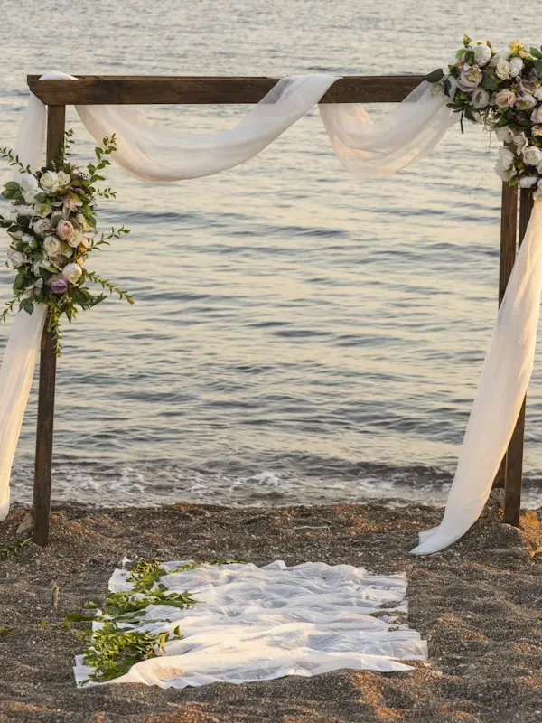Wooden ceremony arch with white drapes and flowers on a beach at sunset